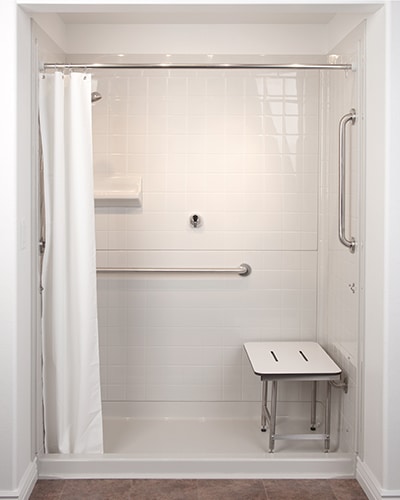 A walk-in shower with heavy weighted white shower curtains, horizontal and vertical grab bars, a wall shelf, a removable bench, and a handheld showerhead holder.
