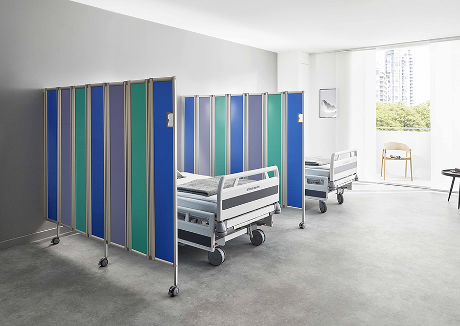 Two hospital beds separated by ropimex® RFW Mobile Folding Screens in a bright, modern hospital room with large windows and minimal furnishings.