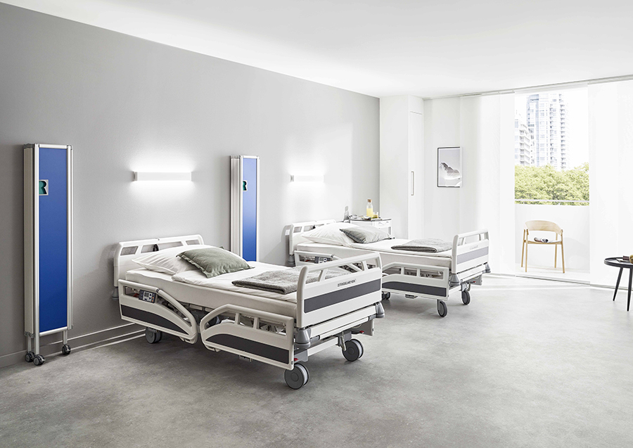 A modern hospital room with two adjustable beds, gray bedding, pillows, and blue ropimex® RFW Mobile Folding Screens. Large windows let in natural light, and there is a chair and small table near the window.