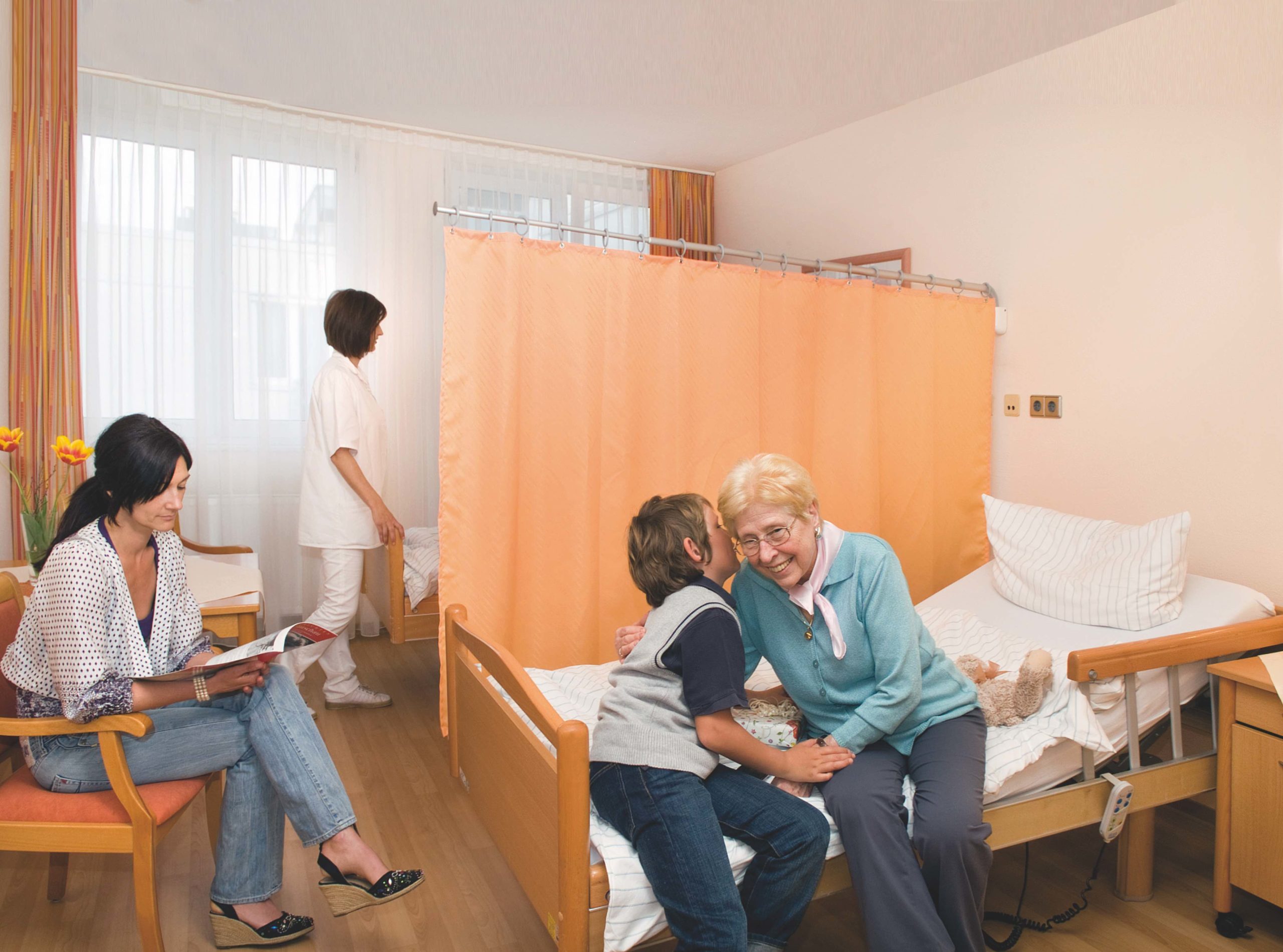 A young boy hugs an elderly woman on a hospital bed, while another woman reads nearby. A nurse walks toward Trevira®CS TCS Privacy Curtains in the bright, clean hospital room.