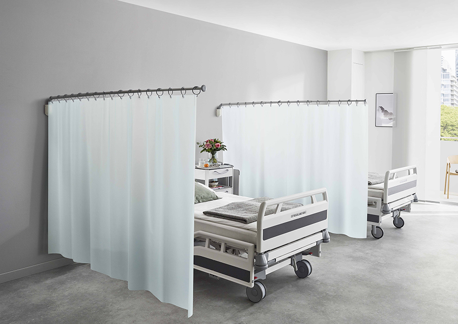 Two hospital beds are separated by Cleanique®CQ Privacy Curtains in a modern room with gray floors, white walls, and natural light from a window. A bouquet of flowers sits on a bedside table.