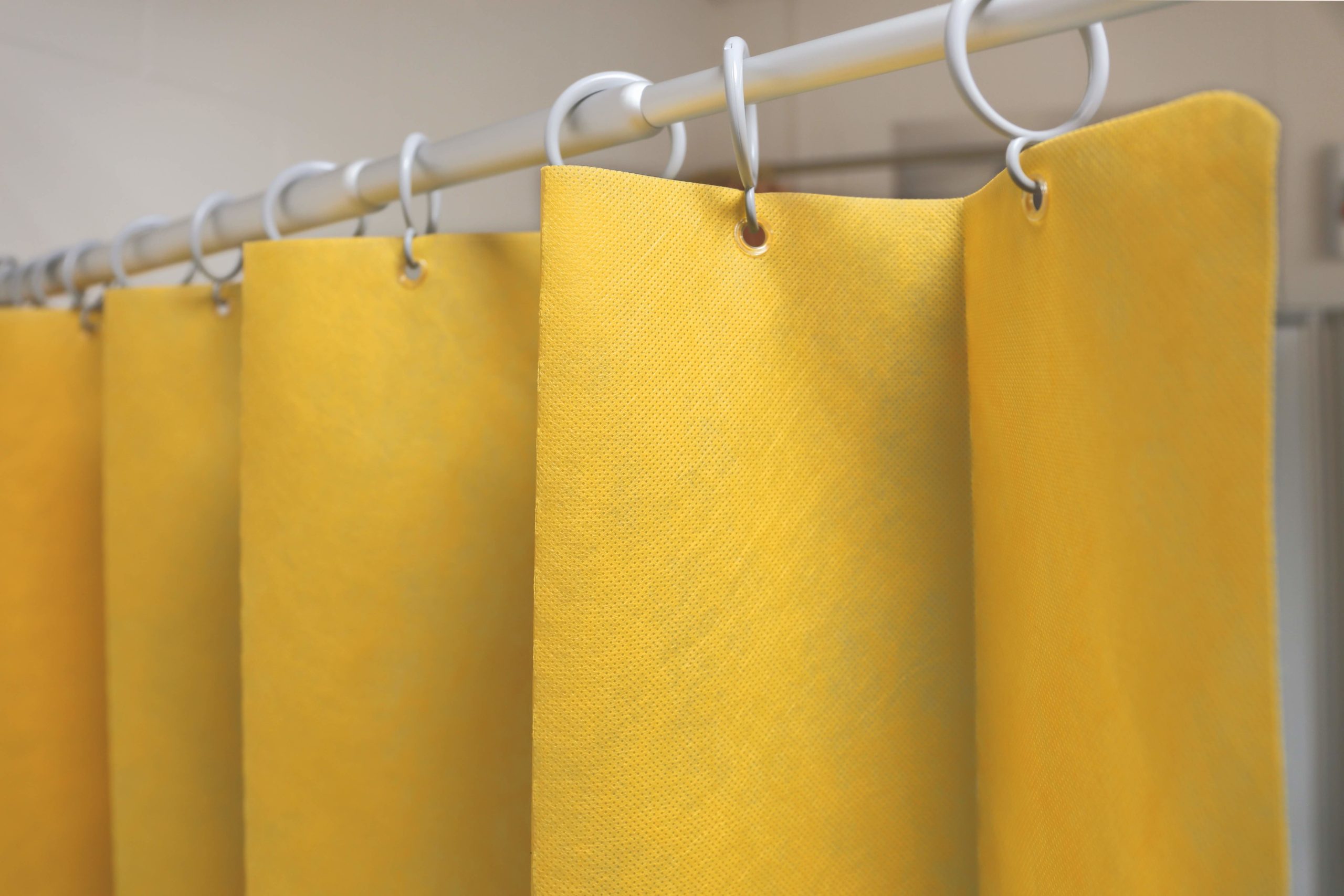 A close-up view of the Zerøtex disposable curtain in yellow hanging from a white rod with plastic hooks in a bathroom.