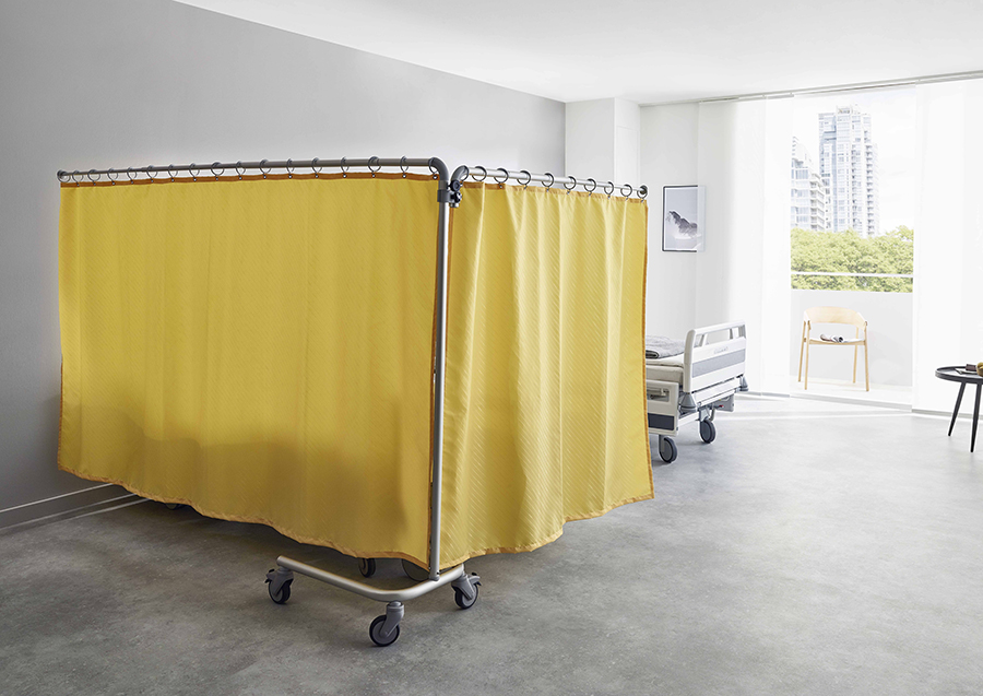A hospital room features a bed partially concealed by a ropimex® FG95 Mobile Privacy Screen on wheels, set beside a large window with city views and a wooden chair placed near the window.