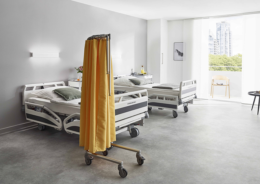 A bright hospital room with two empty beds, ropimex® FG95 Mobile Privacy Screen, a window showing city and trees, a single chair, and a bedside table with personal items.